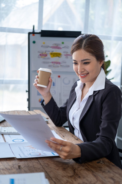 年轻的亚洲快乐美丽的老板商务女性拿着咖啡纸杯，看着纸你的笔记本电脑，微笑的商务女性休息与咖啡图片下载
