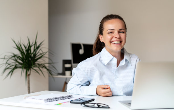 快乐的黑发女人笑着在办公室的办公桌和笔记本电脑前工作。图片下载