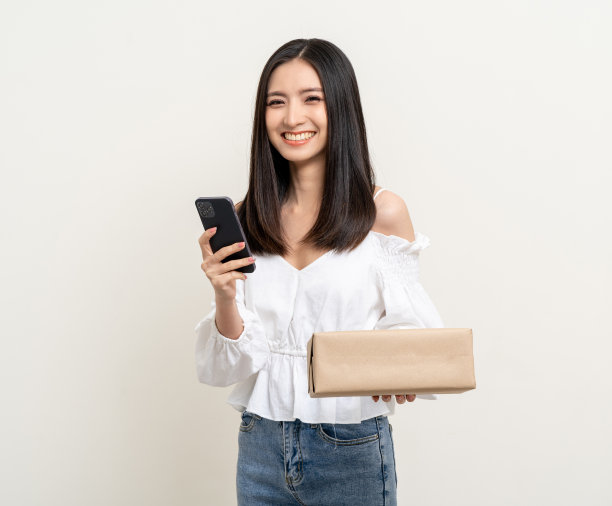 年轻兴奋美丽的亚洲女人拿着智能手机包裹纸板站在孤立的白色背景。从快递服务中接收盒子包裹，支付网上购物应用图片下载