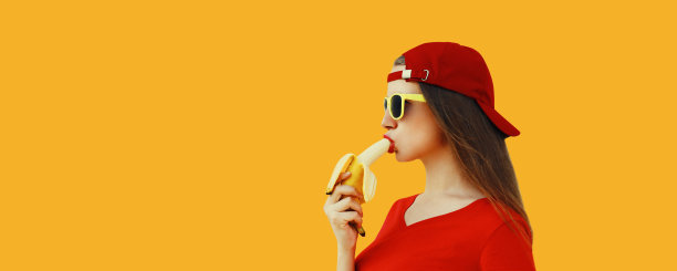 时髦的白人年轻女子戴着橙色背景的红色棒球帽吃香蕉图片下载