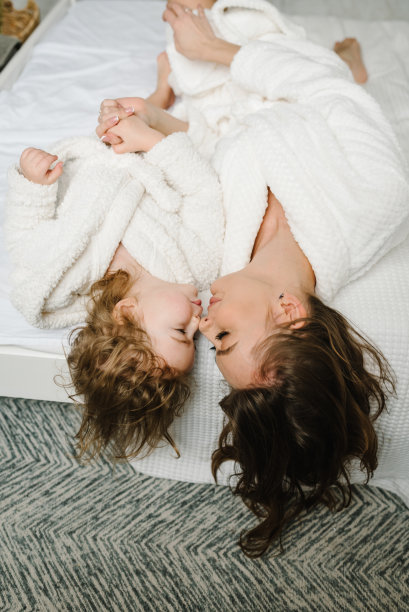 快乐的妈妈和女孩孩子面对面，微笑着。年轻的妈妈带着可爱的小女儿穿着浴衣躺在床上，可爱地看着对方。保健和家庭概念。前视图。美丽的一天。图片下载