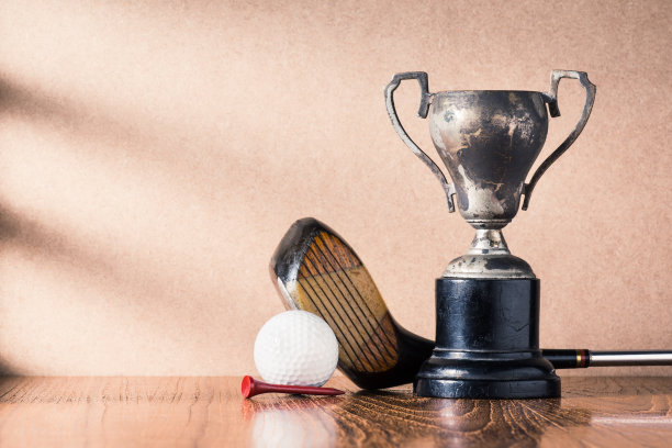 静物摄影:高尔夫球杆(司机)和球与旧奖杯在木桌上对着棕色的墙，左边有窗户灯图片下载