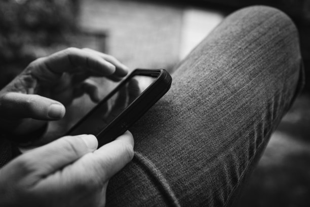 一个穿着休闲牛仔裤坐在户外长凳上看智能手机的人的特写。旅游，联系，放松，工作，在线，电子工作，远程工作概念。黑白复古照片。图片下载