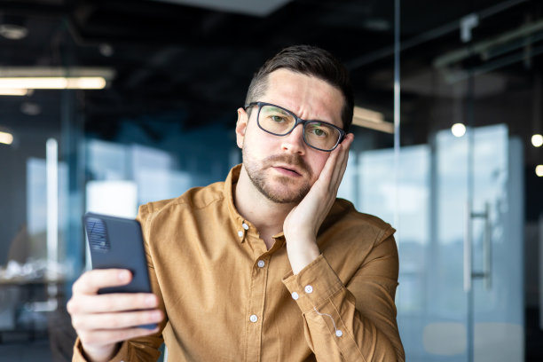 一个心烦意乱的年轻人坐在办公室里，手里拿着电话。在网络游戏中输了钱，通过短信收到坏消息，他忧心忡忡地捂着脸，看着镜头。特写照片图片下载