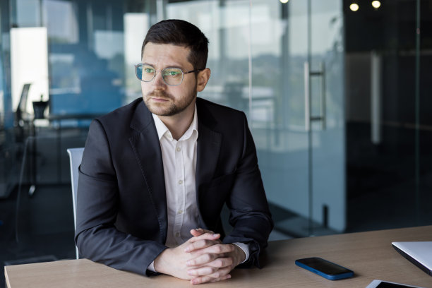 一个严肃的年轻人的肖像，律师，公证人坐在办公室的桌子上，穿着西装，自信地看着一边图片下载
