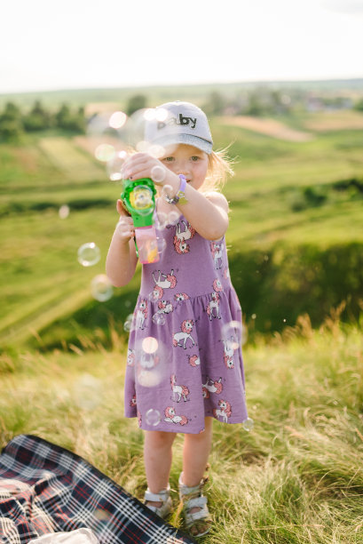 泡沫。快乐的女孩在大自然中吹肥皂泡，抓肥皂泡。夕阳下，孩子走在田野的绿草地上。孩子在山上玩耍，微笑着。图片下载