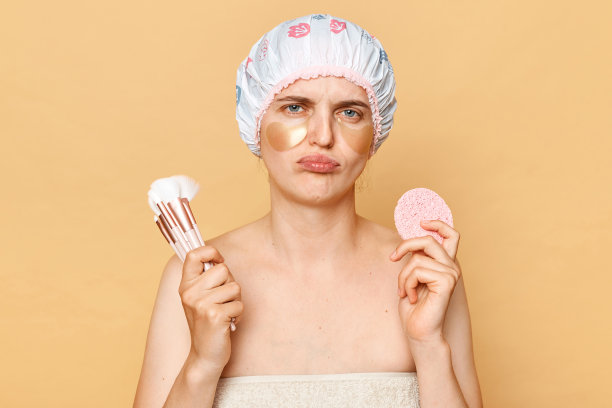 悲伤沮丧的女孩拿着化妆刷和海绵看着镜头，一脸不开心地在早上洗澡后做美容手术，孤立地站在米色的背景下。图片下载