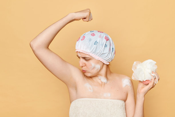 年轻的成年女子抬起双臂，闻着腋下难闻的气味，皱着眉头准备洗澡，拿着海绵，戴着浴帽。图片下载
