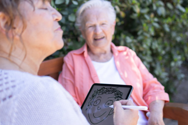 度假时，母亲和女儿微笑着用她的数码平板电脑和电子铅笔画了一幅漫画肖像。趣味和艺术概念。滑稽的场景。鬼混和傻乎乎的鬼脸。图片下载