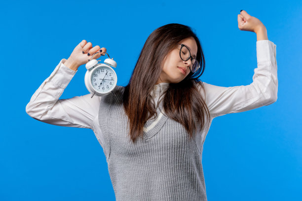 被闹钟叫醒，困倦的女学生手里拿着闹钟。蓝色背景。早上7点。懒惰的青少年没有足够的睡眠，打发时间的观念，教育的生活方式图片下载