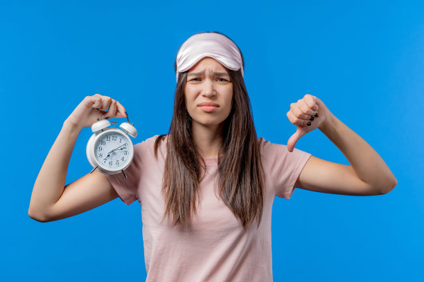 被闹钟叫醒，昏昏欲睡的女人手里拿着闹钟。蓝色背景。早上7点。懒惰的女士没有得到足够的睡眠，打发时间的概念。图片下载