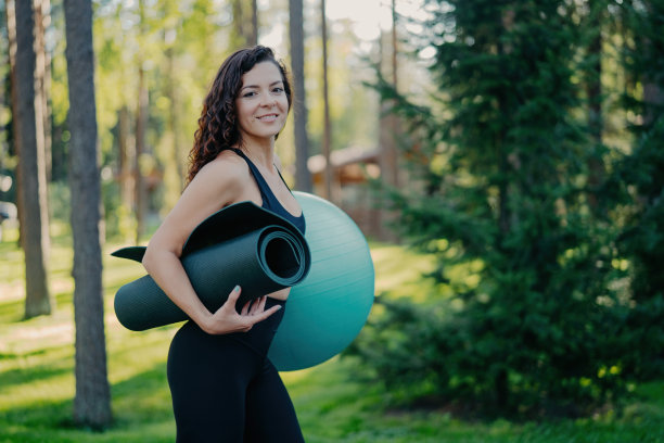 侧面照片中，美丽的卷发黑发女子手持卷起的瑜伽衣和健身球，穿着运动服，在森林或公园里摆姿势，在户外进行健身训练，过着积极的生活方式图片下载