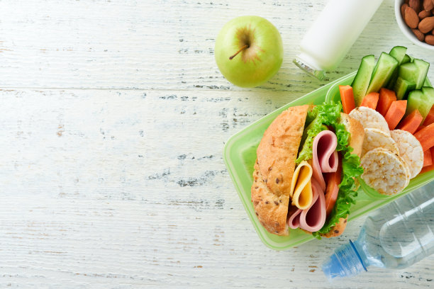 学校午餐的健康食品概念。健康面包，肉三明治与奶酪，苹果，新鲜黄瓜，胡萝卜，坚果在容器，水和酸奶在厨房的背景。回到学校的概念。图片下载