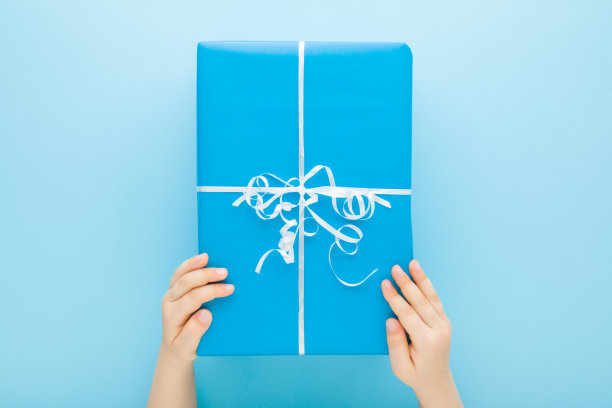 小男孩的手拿着大包装礼盒与白色丝带在浅蓝色的桌子背景。柔和的颜色。特写镜头。祝贺的概念。视角镜头。自顶向下视图。图片下载