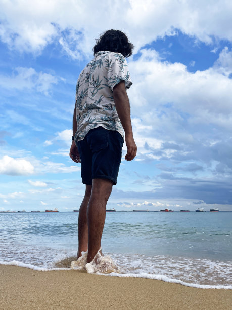 新加坡东公园海滩，印度男游客在度假时站在沙滩上望向大海的特写照片，潮水来了，沙滩上的波浪破碎，晴朗的蓝色多云的天空，后视图图片下载