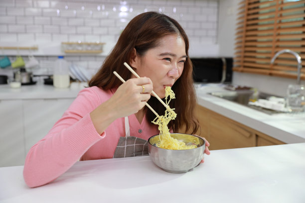 年轻快乐的亚洲卷发女人的肖像坐在桌子上，在厨房里用筷子和砖墙吃着美味的方便面。不健康和廉价食品的生活方式概念图片下载
