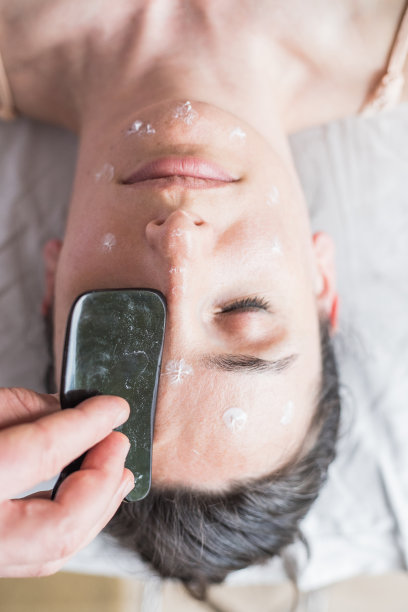 年轻的白人女性正在做面部护理。治疗师正在做刮皮术。中医手法，瓜沙，止痛，治疗，活血。刺激疗法。图片下载