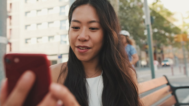 漂亮的黑发女孩穿着白色上衣和牛仔裤，坐在以城市为背景的长凳上微笑着自拍图片下载
