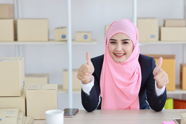 活跃的亚洲女人穿着蓝色的穆斯林套装，坐在电脑旁工作，在线投递包裹。创业小自由职业女孩展示拇指与激动的情绪。图片下载