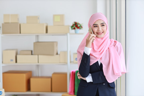 身着蓝色穆斯林套装，头戴粉色帽子的虔诚的亚洲妇女站着，自信地使用手机。女商人站在一旁，拿着包裹投递中小企业的盒子。在家工作概念图片下载