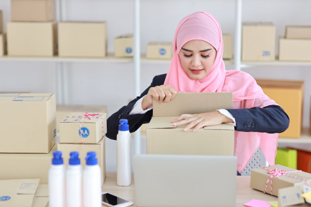 活跃微笑的亚洲女人，穿着蓝色的穆斯林套装，坐在那里从事在线包裹递送工作。创业小公司中小企业自由职业女孩在电脑和手机工作的快乐的脸。图片下载