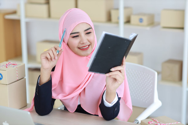 穿着穆斯林套装的活跃亚洲女性坐在那里，用电脑、移动智能手机和在线投递包裹。创业小生意自由职业女孩检查客户订单的书图片下载
