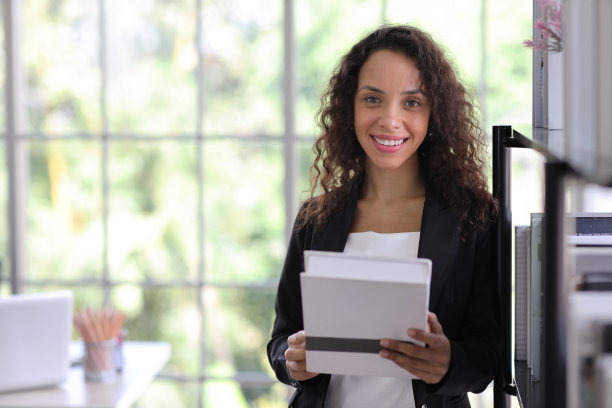 在现代办公室里，自信美丽的年轻非洲女商人站在那里，拿着一本书，带着幸福的微笑看着镜头。成功经营理念图片下载