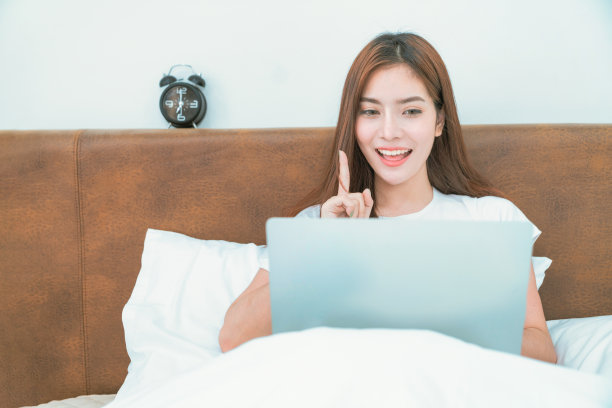 快乐随意的美女坐在家里的床上用笔记本电脑工作。早上，一个快乐的女人躺在床上放松地用笔记本电脑看新闻图片下载