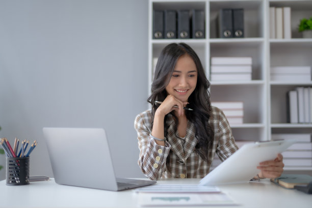 年轻漂亮的女经理或公司职员，拿着会计文件，检查财务数据或营销报告，在办公室用笔记本电脑工作。会计咨询一些文件。图片下载