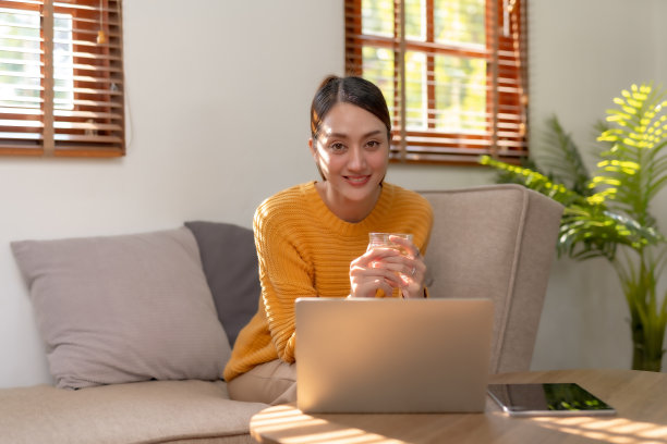 微笑的亚洲女人坐在家里的沙发上使用笔记本电脑。美丽的年轻亚洲女性购物或在社交网络上聊天，玩得开心，看电影，在笔记本电脑项目上自由工作。图片下载