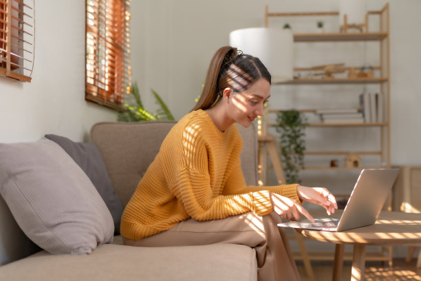 微笑的亚洲女人坐在家里的沙发上使用笔记本电脑。美丽的年轻亚洲女性购物或在社交网络上聊天，玩得开心，看电影，在笔记本电脑项目上自由工作。图片下载