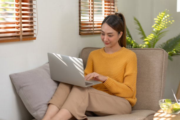 微笑的亚洲女人坐在家里的沙发上使用笔记本电脑。美丽的年轻亚洲女性购物或在社交网络上聊天，玩得开心，看电影，在笔记本电脑项目上自由工作。图片下载