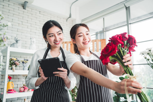 在摆满鲜花和用品的花店里，年轻的花商微笑着在平板电脑上核对订单。图片下载