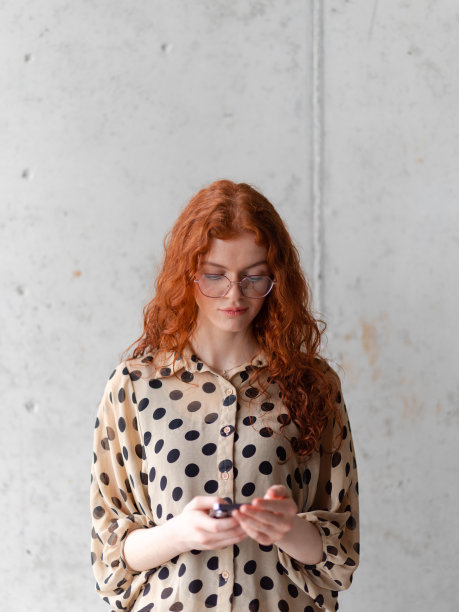 一位有着迷人橙色头发的年轻企业家在时尚的灰色墙壁前摆出自信的姿势，手里拿着智能手机。图片下载