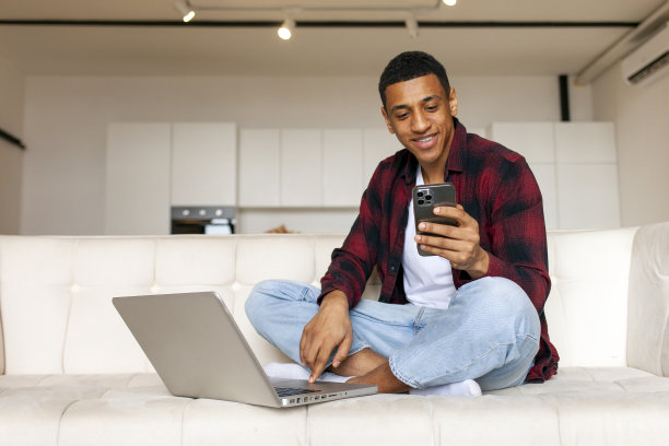 年轻的非洲裔美国人坐在家里的沙发上使用笔记本电脑和智能手机图片下载