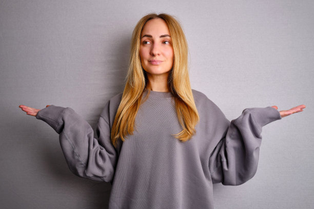 一名身穿灰色运动衫的女子在一面墙旁摆姿势。手势。情感。休闲服。工作室模特在工作。铭文和标志的地方图片下载