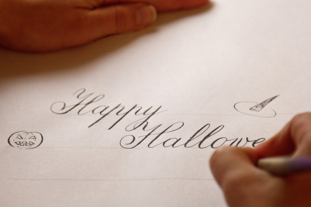 手用墨笔在带条纹的白纸上写下快乐的万圣节。办公桌上的文具近距离俯视图。拼写课和书法练习。模板，布局，背景图片下载