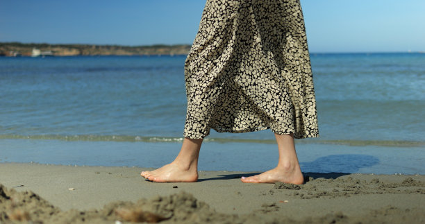 海边沙滩上，女性赤脚站在水里的特写镜头。在海岸线漫步时穿着长裙的女人的腿。图片下载