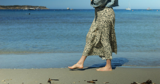 不知名的时尚女子在海滨长廊的沙滩上缓慢优雅地迈步。夏日的微风中，女学生赤脚走在海岸线上。图片下载