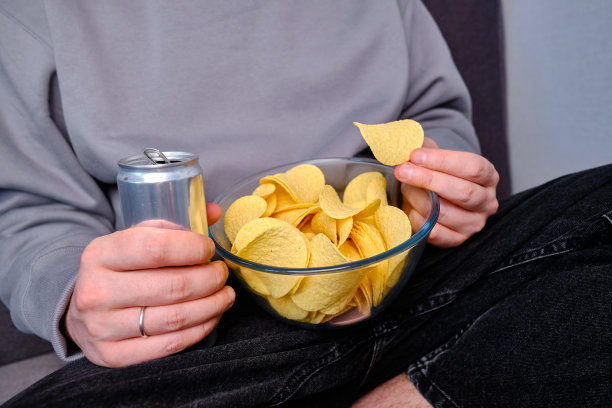 一个男人在沙发上用一个透明的碗吃酥脆的薯片。快餐。卡路里和饮食。垃圾食品图片下载
