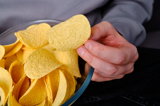 一个男人在沙发上用一个透明的碗吃酥脆的薯片。快餐。卡路里和饮食。垃圾食品图片下载