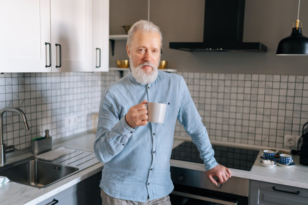 英俊的灰色头发成熟的成年男子在家里喝咖啡，早上看着相机的肖像。思来想去的老人喝着茶，沉浸在沉思中图片下载