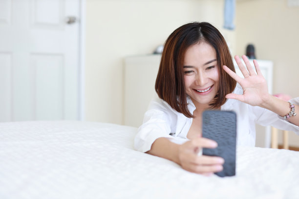 年轻迷人的亚洲女人穿着白衬衫，用VDO从手机打电话，坐在卧室里跟别人打招呼图片下载