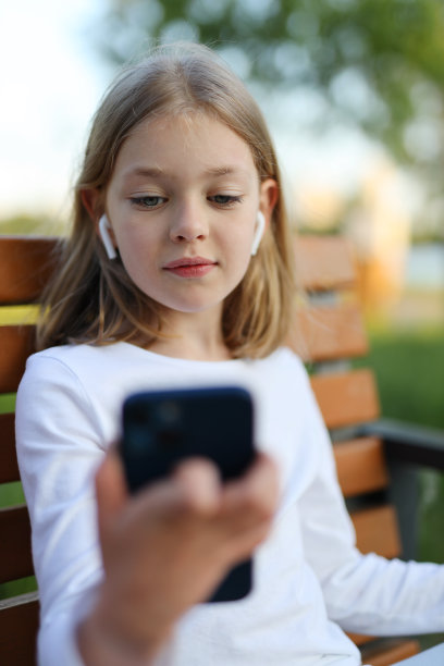 一个小女孩拿着智能手机和耳机坐在公园的长椅上。图片下载