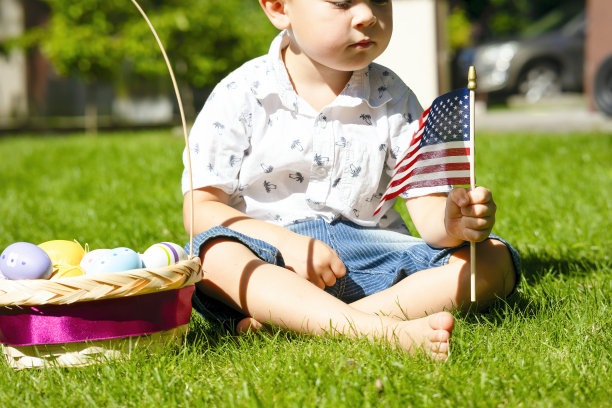 一个孩子坐在草地上，手里拿着一面美国国旗，旁边放着一篮子复活节彩蛋。图片下载