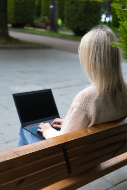 一个年轻的金发女子的后视图，膝上放着一台笔记本电脑，坐在公园的长椅上。女孩在公园里用笔记本电脑打字。垂直图图片下载