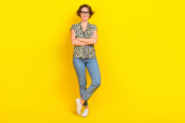 自信的商务女士抱着双臂，穿着眼镜印花衬衫牛仔裤鞋子孤立的黄色背景图片下载
