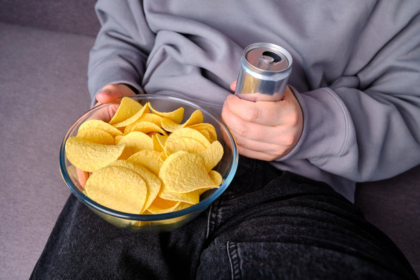 一个男人在沙发上用一个透明的碗吃酥脆的薯片。快餐。卡路里和饮食。垃圾食品图片下载