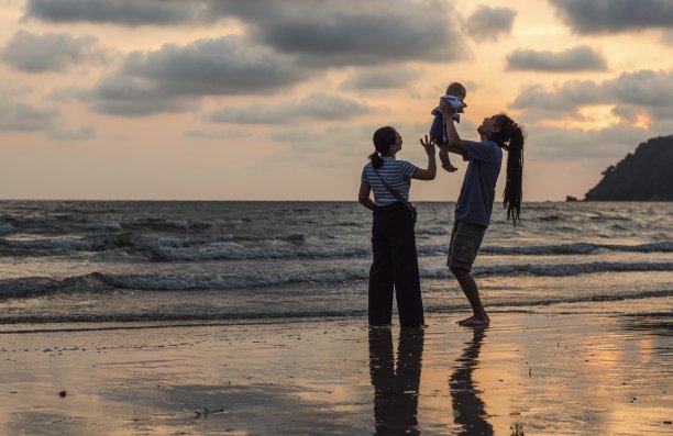 假日，快乐的一家人在海滩上旅行的剪影，父亲在黑色的沙滩上，沿着日落的海边冲浪，愉快地把女婴吐出来。积极的家长和人们在夏天的户外活动。图片下载