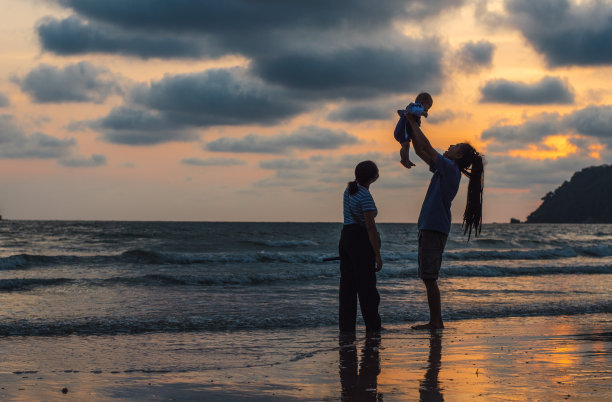 假日，快乐的一家人在海滩上旅行的剪影，父亲在黑色的沙滩上，沿着日落的海边冲浪，愉快地把女婴吐出来。积极的家长和人们在夏天的户外活动。图片下载